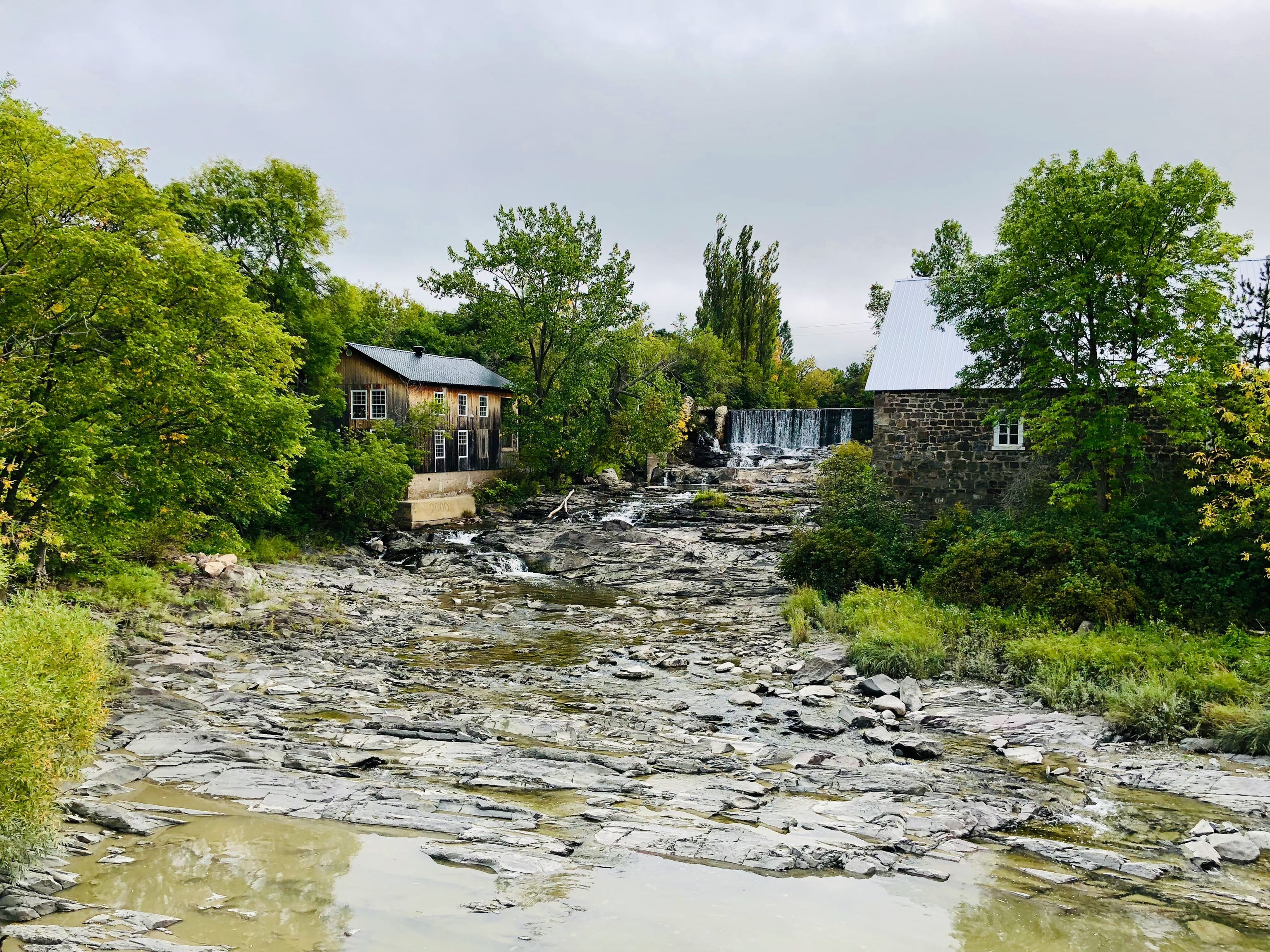 Vue d'un lit de rivière rocailleux avec peu d'eau, entouré de forêts, de bâtiments anciens et d'une petite chute d'eau.