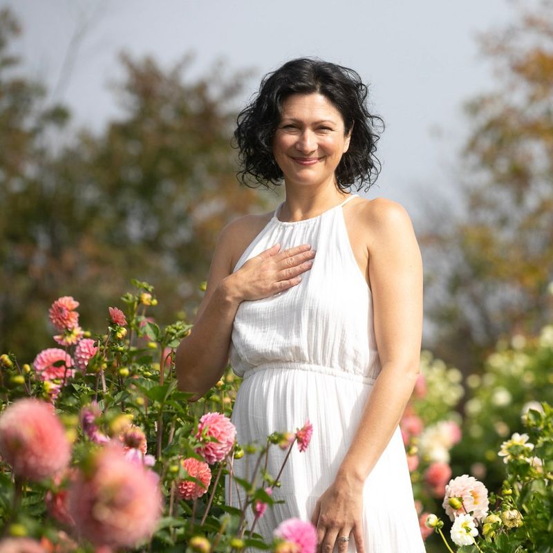 Christine Dion en robe blanche, la main sur le cœur, parmi les dahlias roses.